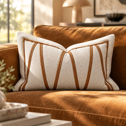 A lumbar decorative throw pillow with a textured cream linen fabric and vertical brown embroidered stripes, styled on a warm rust-orange velvet sofa in a sunlit living room.8