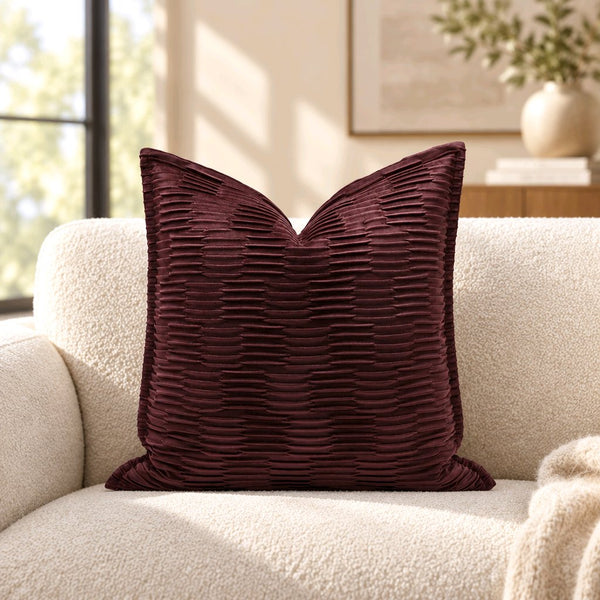 A square velvet throw pillow in deep burgundy with a textured, pleated horizontal ribbed pattern, resting on a cream boucle sofa in a brightly lit living room.2